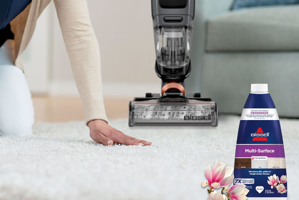 Płyn do odkurzaczy myjących BISSELL CrossWave Max 2765N/Pet Pro 2225N/X7 Plus Pet Pro 3400N / SpinWave 20522 1000 ml Odkurzacz pionowy Bissell CrossWave na puszystym białym dywanie obok butelki płynu Multi-Surface Spring Breeze i kwiatów magnolii.