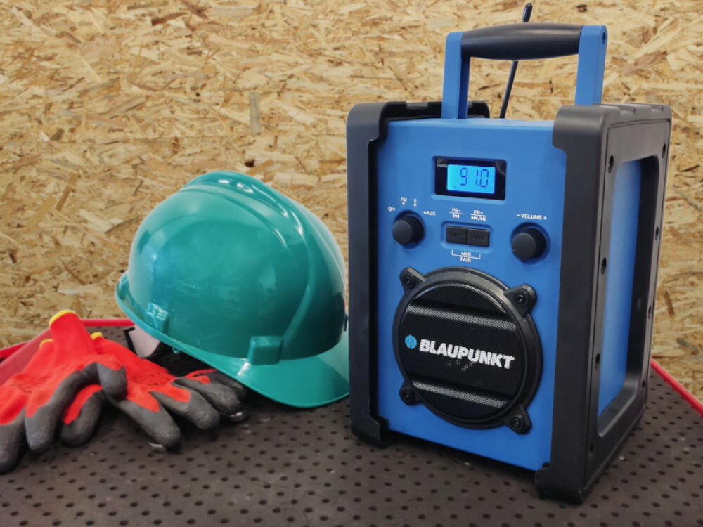 Radio budowlane BLAUPUNKT PP30BT JOBSITE, uchwyt, waga, wymiar, odporność, wytrzymałość, Radio stoi na blacie wraz z kaskiem budowlanym i rękawicami, w tle ściana z płyty OSB