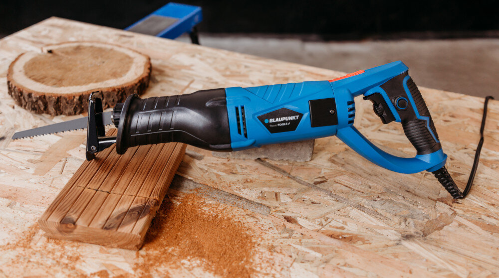 Na stole warsztatowym leży niebiesko-czarna piła szablasta z widocznym logo \'BLAUPUNKT RS6010\'. Obok piły znajduje się kawałek drewna z trocinami.