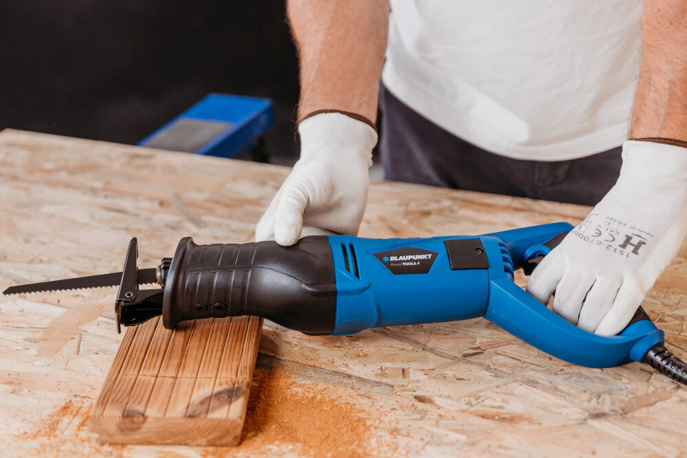 Osoba w białych rękawicach trzyma niebiesko-czarną piłę szablastą marki BLAUPUNKT RS6010, która jest umieszczona na drewnianej desce. Na stole widoczne są trociny.