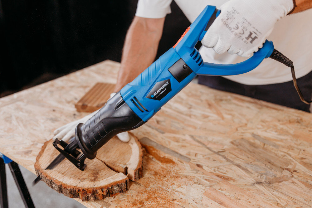 Osoba trzyma niebieską piłę szablastą, która przecina kawałek drewna na stole warsztatowym. Na piłę widoczne jest logo \'BLAUPUNKT\'.