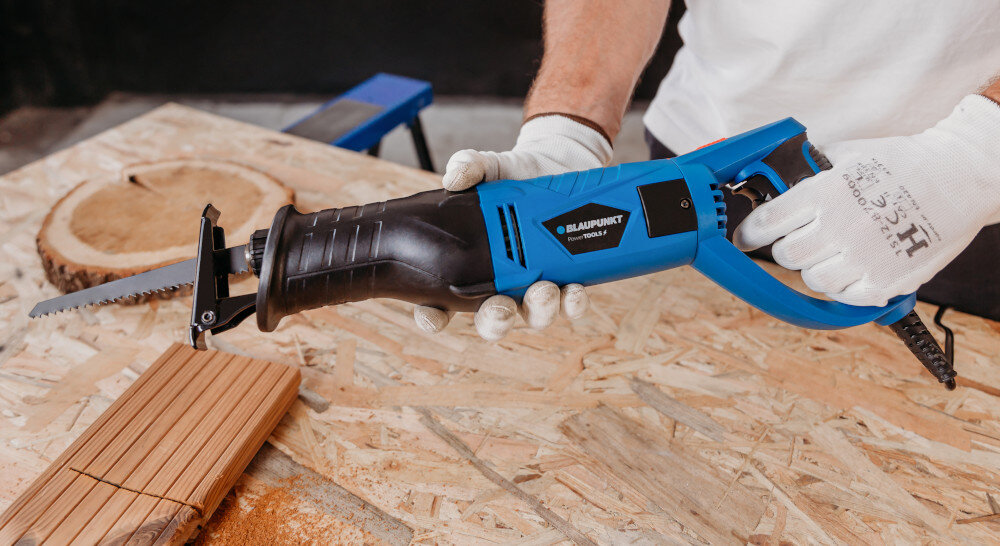 Osoba trzyma piłę szablastą w kolorze niebiesko-czarnym na stole warsztatowym. Na piłę widoczne jest logo \'BLAUPUNKT\' oraz napis \'RS6010\'.