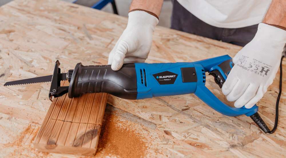 Osoba w rękawicach trzyma niebiesko-czarną piłę szablastą marki BLAUPUNKT, która przecina drewnianą deskę na stole roboczym. Na piłę padają trociny, a na obudowie widoczne jest logo \'BLAUPUNKT Power Tools\'.