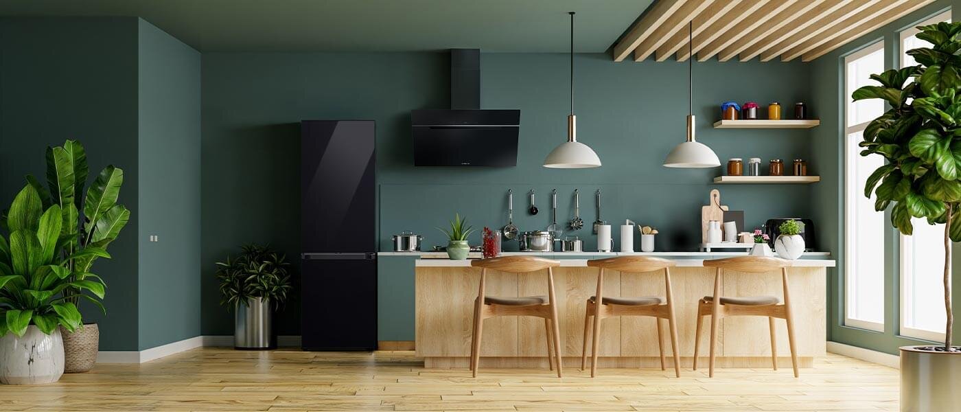 Nowoczesna kuchnia z ciemnozielonymi ścianami, drewnianą podłogą i wyspą kuchenną z czterema krzesłami. Po lewej stronie stoi czarna lodówka, a na blacie znajdują się różne akcesoria kuchenne.