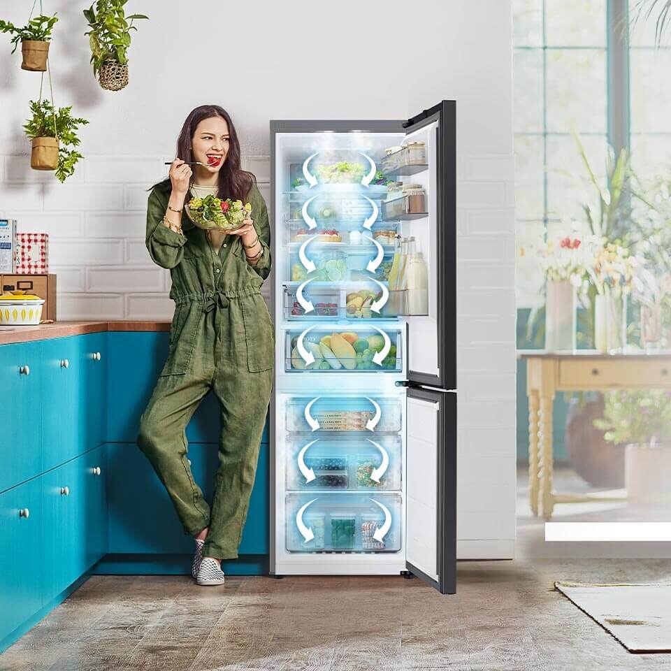 Grafika przedstawia, jak w lodówce Samsung RB34C7B5D22 z oferty Media Expert dzięki All-Around Cooling chłodne powietrze dociera do każdego miejsca w chłodziarce i zamrażarce Kobieta w zielonym kombinezonie stoi obok otwartej lodówki, jedząc sałatkę. Wnętrze lodówki jest dobrze oświetlone i wypełnione różnymi produktami spożywczymi.