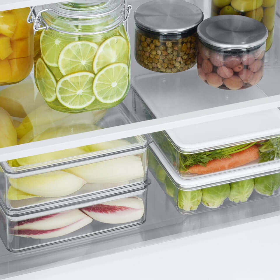 Wnętrze lodówki z różnymi produktami spożywczymi w przezroczystych pojemnikach, w tym plasterki limonki w słoiku, marchewki, brukselka i inne warzywa. Na górnej półce znajdują się również dwa słoiki z metalowymi pokrywkami, zawierające różne produkty.