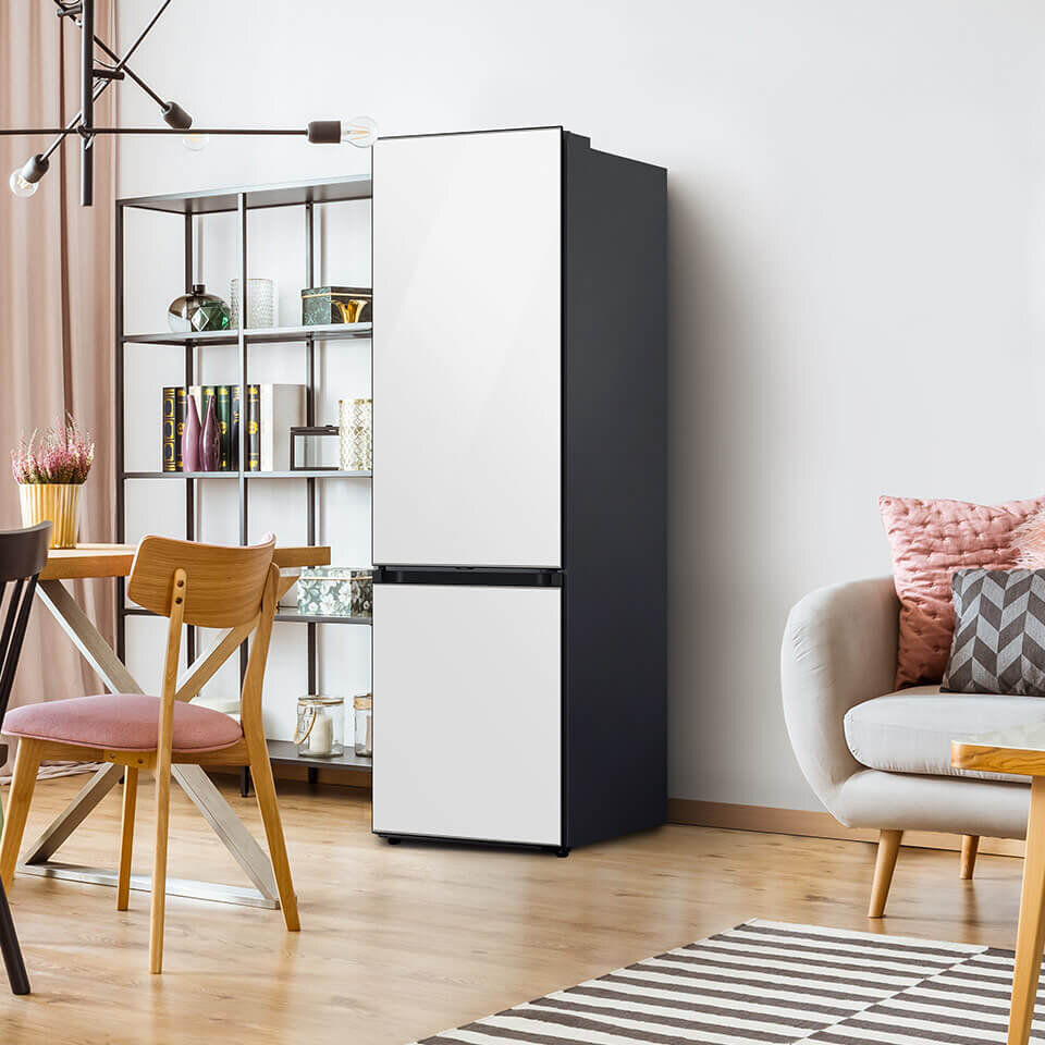 Wnętrze nowoczesnego salonu z lodówką o białych drzwiach i czarnych bokach. Obok znajduje się stół z krzesłami oraz regał z książkami i dekoracjami.