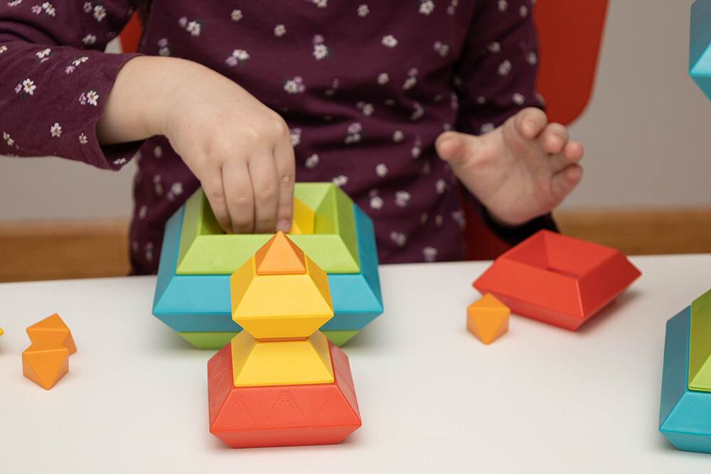 Zbliżenie na dłonie dziecka układającego żółty element wewnątrz błękitnej formy klocków edukacyjnych na białym tle.