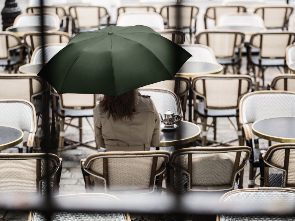 Parasolka LEXON Mini Hook LU21LB1 Jasnoniebieski osoba siedzi na krześle zasłonięta czarnym parasolem funkcja automatycznego rozkładania jedno kliknięcie ochrona przed deszczem lub słońcem średnica (122 cm) wygodna osłona na codzienne spacery w drodze do pracy lub szkoły solidna konstrukcja trwałość komfort użytkowania w każdej sytuacji 