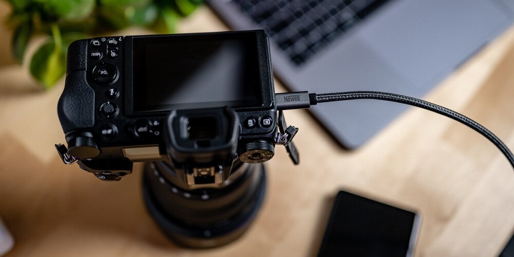 Szary kabel USB w materiałowym oplocie wpięty w korpus aparatu fotograficznego, w tle rozmyte biurko i roślina