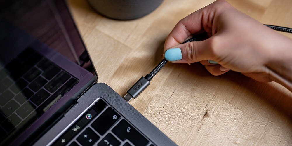 Dłoń podłącza pleciony kabel USB do laptopa leżącego na drewnianym biurku, widoczna klawiatura i złącze