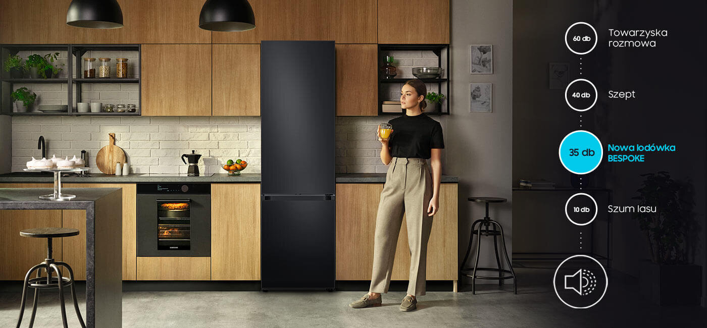 Kobieta stoi w kuchni obok grafitowej lodówki, trzymając szklankę z napojem. Obok znajduje się diagram z poziomami hałasu: 60 dB Towarzyska rozmowa, 40 dB Szept, 35 dB Nowa lodówka BESPOKE, 10 dB Szum lasu.