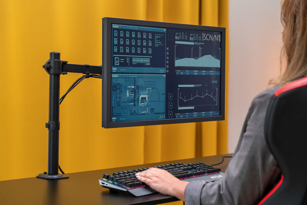 Uchwyt XQUANTUM MA10341 Kobieta siedząca przed biurkiem, na którym zamontowany jest monitor na czarnym uchwycie biurkowym. Monitor jest ustawiony w orientacji poziomej i wyświetla interfejs z danymi i wykresami. Kobieta pisze na klawiaturze, a w tle widać żółtą zasłonę.