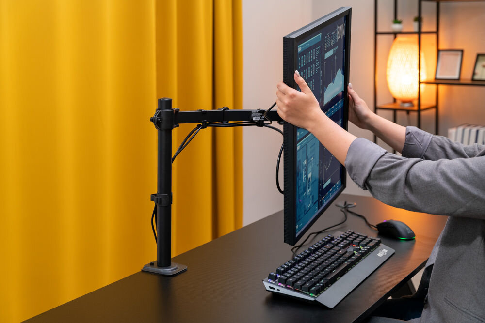Uchwyt XQUANTUM MA10341 Widok z boku na biurko, gdzie kobieta w szarej marynarce oburącz trzyma monitor zamontowany na czarnym uchwycie, dostosowując jego pozycję. Monitor jest w orientacji pionowej i wyświetla interfejs z danymi. W tle widać żółtą zasłonę i stojącą lampę.