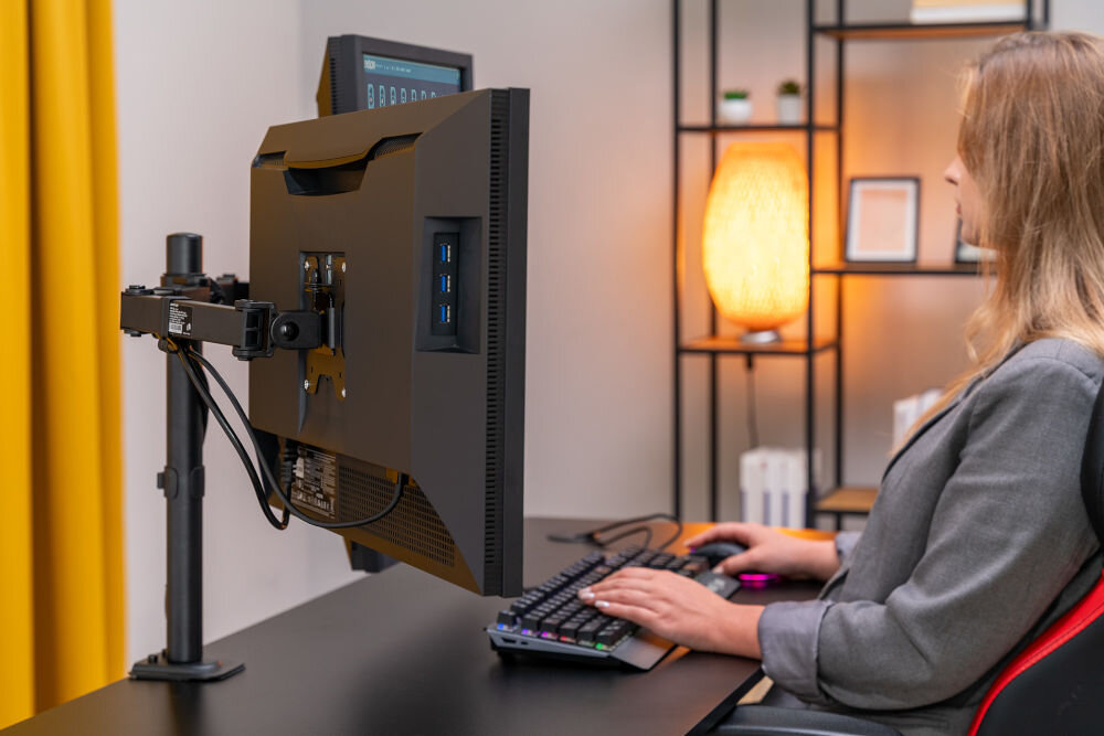 Uchwyt XQUANTUM MA10322 Monitor zamontowany na czarnym uchwycie biurkowym xquantum ustawiony w pozycji poziomej nad blatem. Osoba siedząca przy biurku pracuje na klawiaturze, a w tle widoczna jest żółta zasłona.