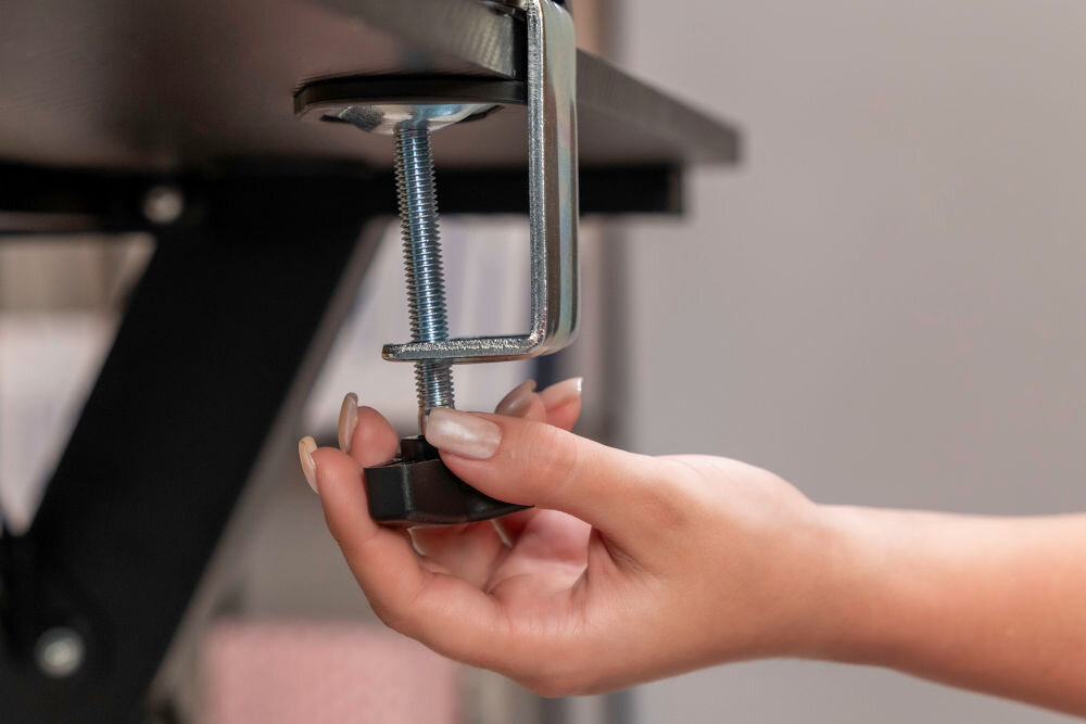 Uchwyt XQUANTUM MA10322 Zbliżenie na dłoń dokręcającą czarną gałkę dolnego zacisku uchwytu monitora xquantum. Metalowa klamra typu C jest stabilnie przymocowana do krawędzi czarnego blatu biurka.