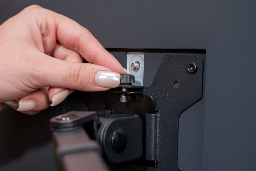 Uchwyt XQUANTUM MA10322 Zbliżenie na tył monitora, gdzie dłoń reguluje małe pokrętło przy płytce VESA uchwytu xquantum. Zdjęcie prezentuje system mocowania i regulacji kąta nachylenia ekranu.
