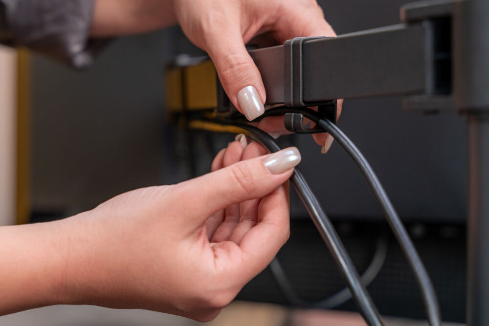 Uchwyt XQUANTUM MA10322 Zbliżenie na tylne ramię uchwytu monitora xquantum, na którym dłonie układają czarne kable w specjalnym klipsie do zarządzania przewodami.