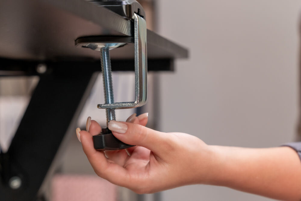 Osoba montuje uchwyt do monitora przy pomocy metalowego zacisku z gwintem. Uchwyt jest przymocowany do krawędzi blatu.