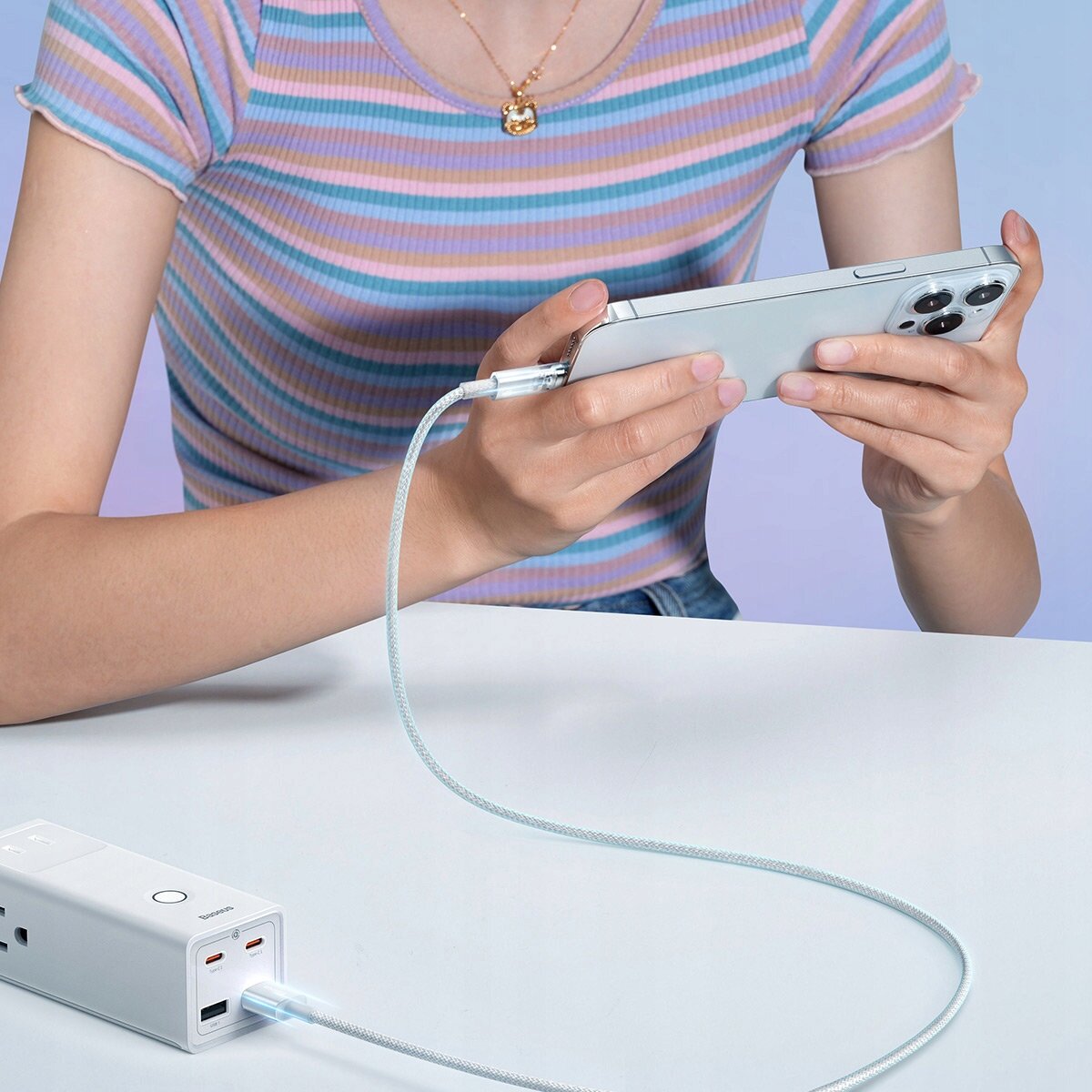 Osoba trzyma smartfon podłączony do białego powerbanku za pomocą plecionego kabla. Na powerbanku widoczne są dwa porty USB i logo \'Baseus\'.