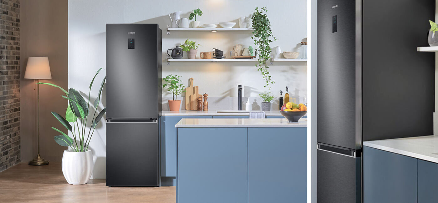 Grafitowa lodówka z logo SAMSUNG stoi w nowoczesnej kuchni z niebieskimi szafkami i białym blatem. Na półkach znajdują się rośliny i naczynia, a obok lodówki stoi lampa i roślina w doniczce.