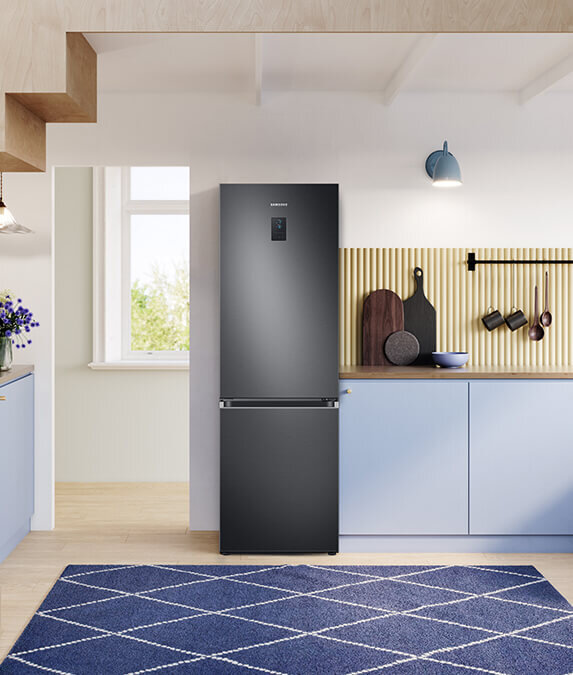 Grafitowa lodówka z logo \'SAMSUNG\' stoi w nowoczesnej kuchni z jasnymi szafkami i drewnianym blatem. Na blacie znajdują się deski do krojenia i miska, a na ścianie wisi lampa i kilka przyborów kuchennych.