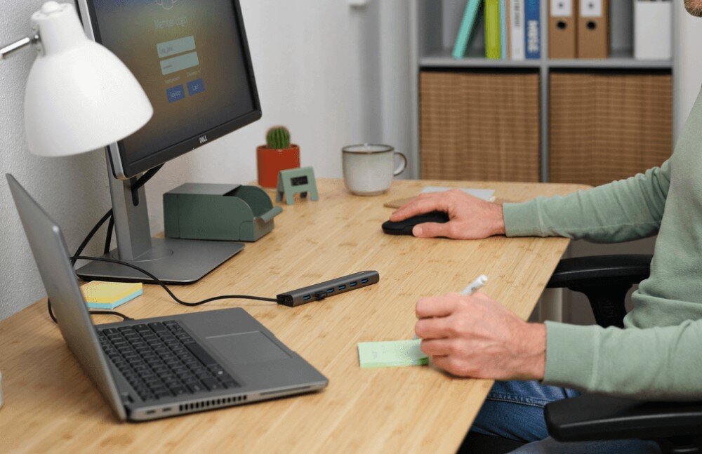 Hub TRUST Halyx 24967 Osoba przy biurku pracująca z notatkami i myszką obok laptopa podłączonego do długiego szarego rozdzielacza portów USB