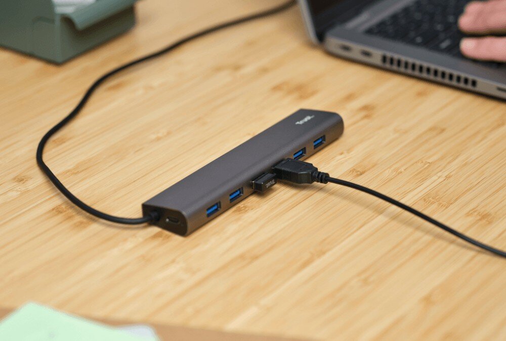 Hub TRUST Halyx 24967 Szary hub USB podłączony długim kablem do portu laptopa leżący na drewnianym biurku obok dłoni w miętowym swetrze
