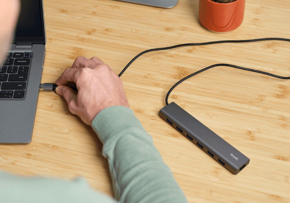 Hub TRUST Halyx 24967 Dłoń wpinająca mały czarny adapter do szarego huba USB leżącego na biurku przy laptopie