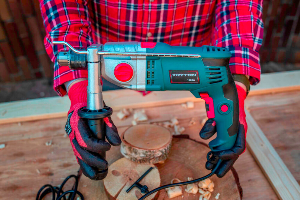 Wiertarka udarowa TRYTON TDW1050A pomieszczenie drewniany blat roboczy osoba trzyma wiertarkę w dłoniach do wiercenia w drewnie stali i betonie moc 1050 W funkcja udaru otwory w twardych materiałach o średnicy do 13 mm niewielka budowa niska waga