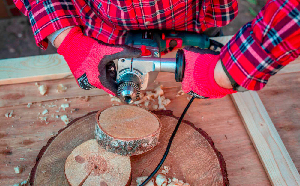 Wiertarka udarowa TRYTON TDW1050A pomieszczenie drewniany blat roboczy osoba trzyma wiertarkę w dłoniach drewniane elementy dodatkowy uchwyt boczny zwiększa stabilność prowadzenia wiertarki poprawia bezpieczeństwo pracy główna rękojeść ma ergonomiczną konstrukcję antypoślizgową powłoką lepsza kontrola