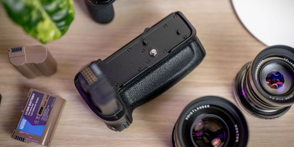 Na drewnianym blacie znajduje się uchwyt do aparatu, obok którego leżą dwa obiektywy i bateria. Na baterii widoczny jest tekst: \'NEWELL Battery Pack\'.
