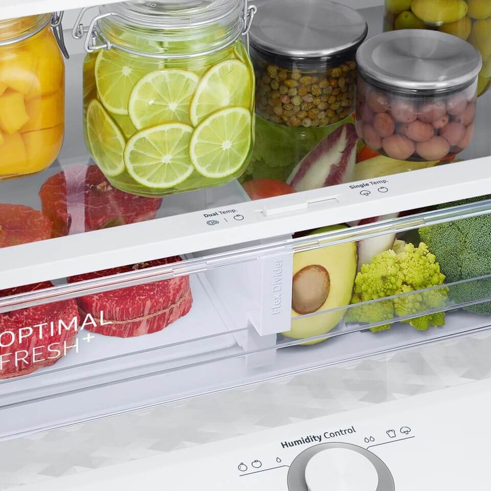 Wnętrze lodówki z widocznymi szklanymi pojemnikami na żywność, wypełnionymi plasterkami limonki, kawałkami mango i innymi produktami. Na szufladzie widoczny jest napis \'OPTIMAL FRESH+\', a poniżej znajduje się pokrętło \'Humidity Control\'.