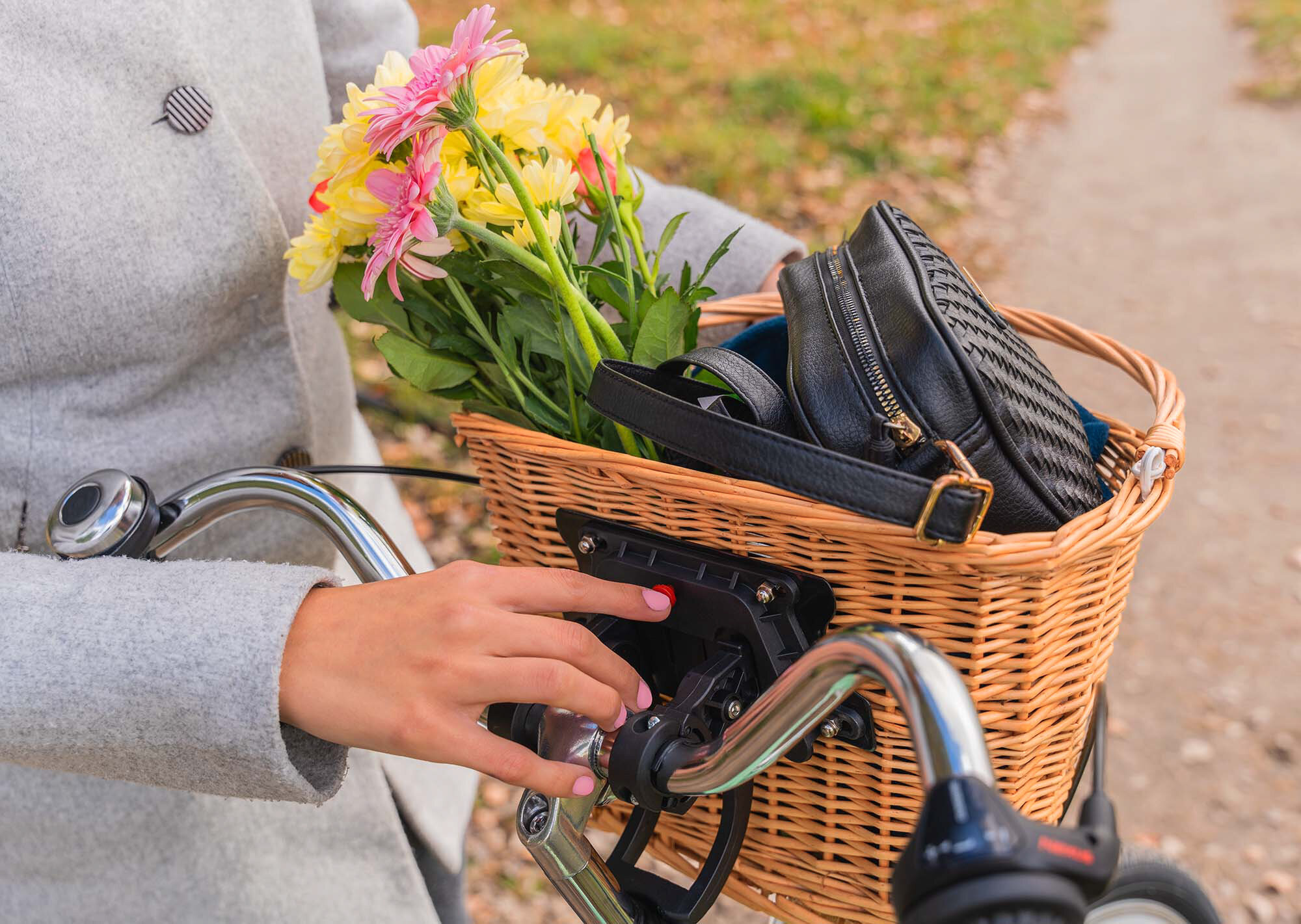 Koszyk wiklinowy na rower S-LINE VKS-502 Zbliżenie na dłoń kobiety w szarym płaszczu, wsuwającą wiklinowy kosz na adapter szybkiego montażu przy kierownicy roweru. Kosz wypełniony kwiatami i torebką. Pokaz mechanizmu klik w trakcie użycia.