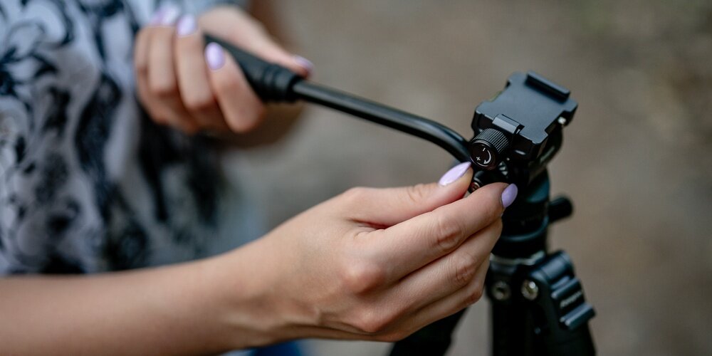 Zbliżenie na dłonie kobiety regulującej głowicę statywu fotograficznego Fotopro; uchwyt ustawiany precyzyjnie w terenie.
