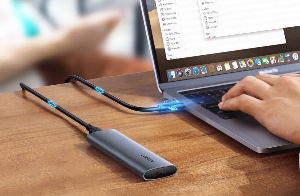 Obudowa dysku UGREEN CM400 10902 Przenośna obudowa na dysk podłączona czarnym kablem do bocznego portu laptopa stojącego na drewnianym blacie, na którym widoczna jest również dłoń obsługująca gładzik