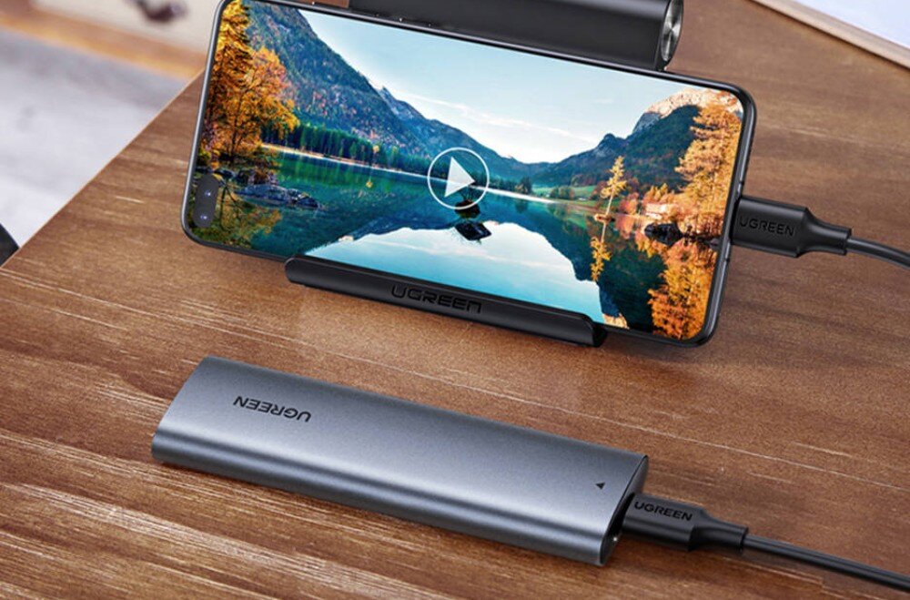 Obudowa dysku UGREEN CM400 10902 Urządzenie zewnętrzne podpięte kablem do smartfona opartego na podstawce, na którego ekranie wyświetlany jest film z górskim krajobrazem