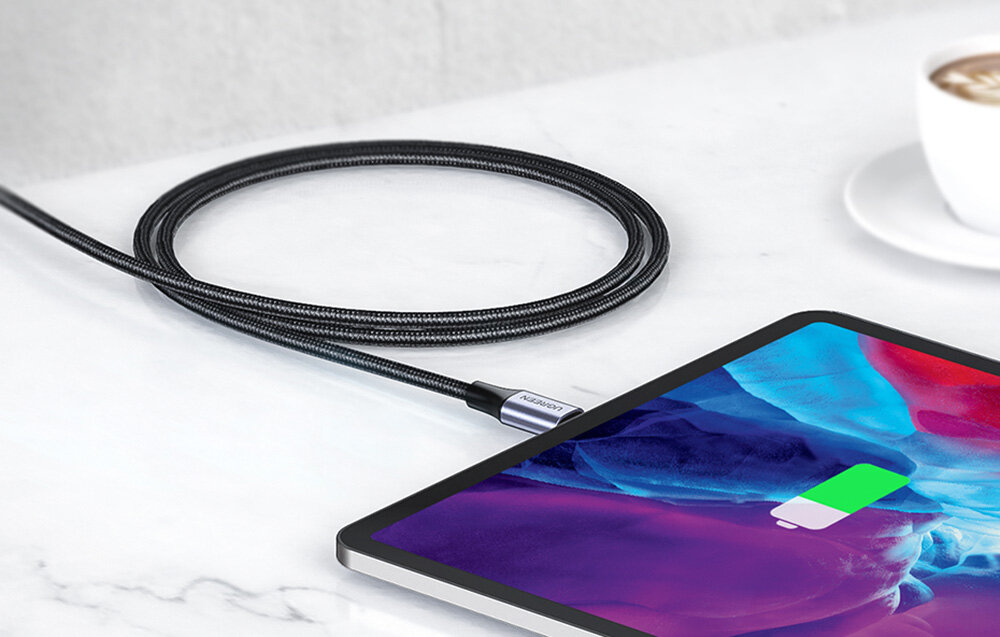 Na białym stole leży czarny kabel podłączony do tabletu, który pokazuje ikonę ładowania. Na końcówce kabla widoczny jest napis \'UGREEN\'.
