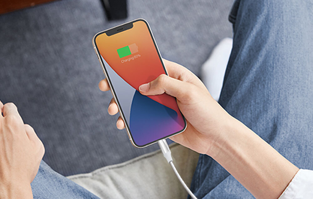 Osoba trzyma smartfon z widocznym ekranem, na którym jest ikona ładowania baterii. Telefon jest podłączony do białego kabla USB-C do Lightning.