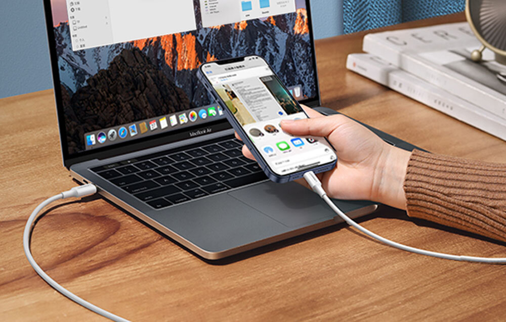 Osoba trzyma smartfon podłączony do laptopa za pomocą białego kabla USB-C do Lightning. Na ekranie laptopa widoczne są otwarte okna aplikacji.