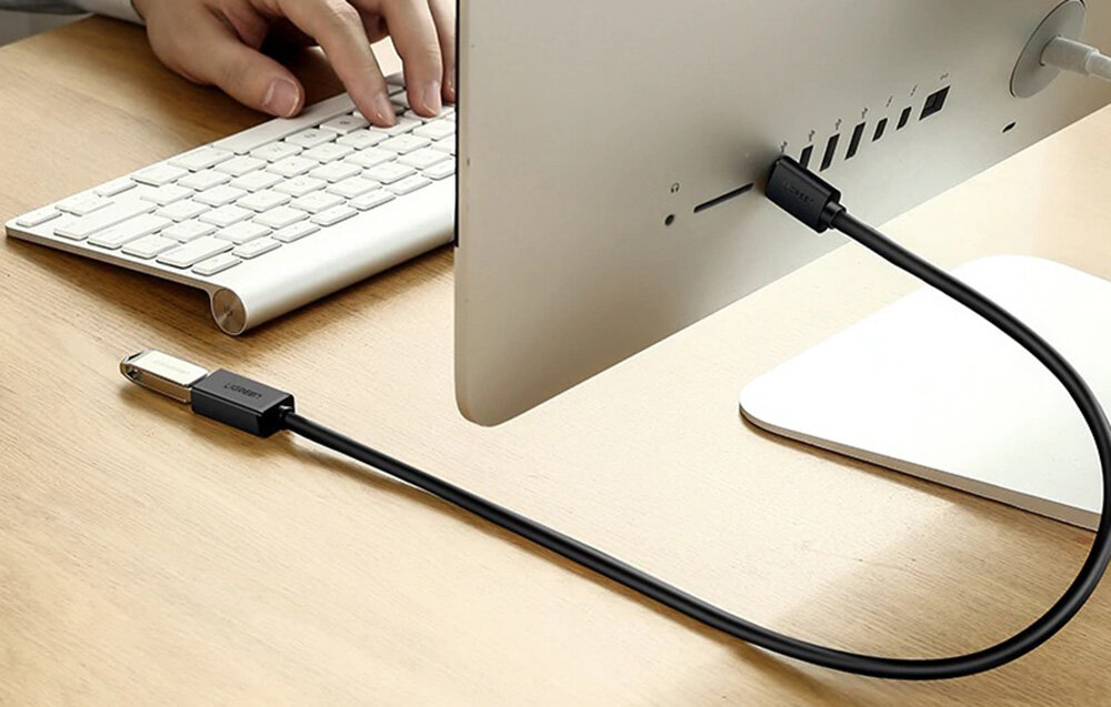 Czarny kabel USB 3.0 podłączony do komputera stacjonarnego, z widocznym logo \'UGREEN\' na wtyczce. Obok znajduje się klawiatura i ręce osoby piszącej na niej.