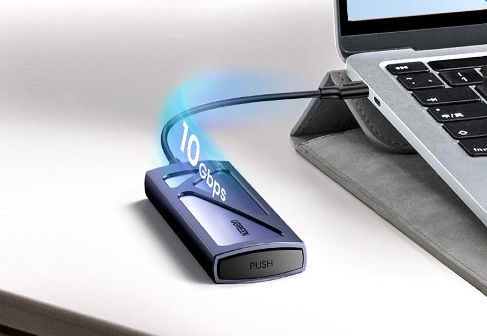 Obudowa dysku UGREEN 15512 Zbliżenie na obudowę dysku zewnętrznego leżącą obok laptopa. Nad kablem łączącym urządzenia widnieje cyfrowo dodany napis '10 Gbps' oznaczający prędkość przesyłu danych