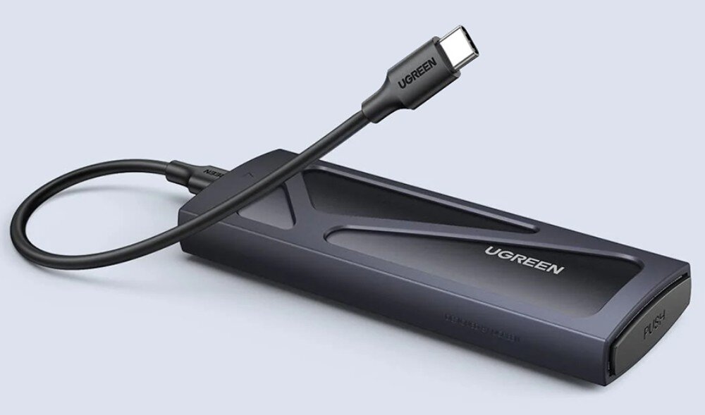 Obudowa dysku UGREEN 15512 Perspektywiczny widok na ciemnoszarą obudowę dysku z wpiętym krótkim kablem zakończonym wtyczką USB-C. Na jednym z końców urządzenia znajduje się przycisk z napisem 'PUSH'