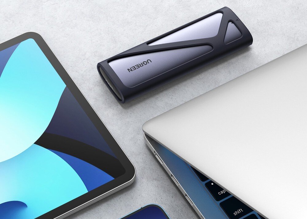 Obudowa dysku UGREEN CM400 Przenośna obudowa na dysk leżąca na jasnym blacie obok tabletu i częściowo otwartego laptopa