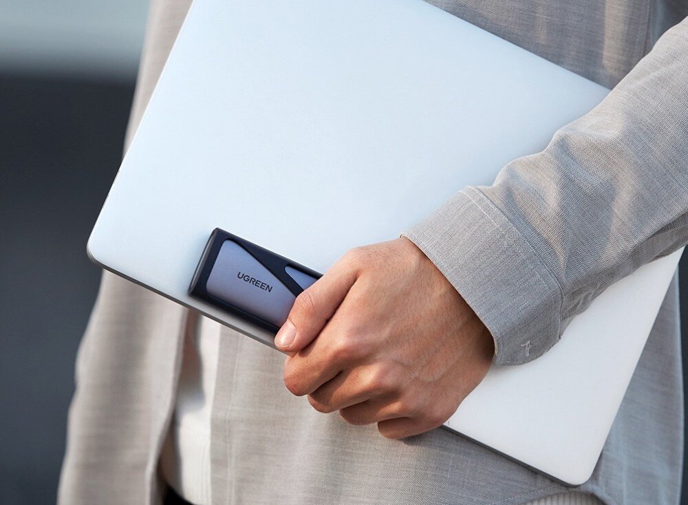 Obudowa dysku UGREEN CM400 Osoba trzymająca pod pachą srebrnego laptopa oraz obudowę na dysk w dłoni