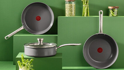 Na zielonym tle znajdują się trzy patelnie z metalowymi uchwytami, z czerwonym okręgiem na środku powierzchni. W tle widoczne są słoiki z produktami spożywczymi i zielone warzywa.