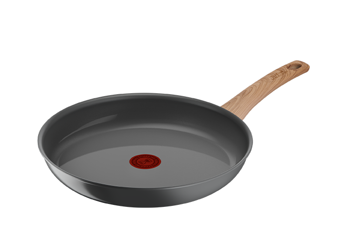 Na obrazie widoczna jest patelnia z ciemnym wnętrzem i drewnianą rączką. Na środku patelni znajduje się czerwony okrągły symbol.