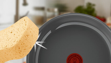 Na obrazie widoczna jest szara patelnia z czerwonym logo Thermo Signal oraz żółta gąbka zbliżająca się do jej powierzchni.