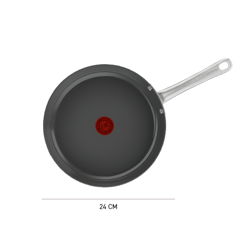 Na obrazie widoczna jest patelnia z ciemnym wnętrzem i metalowym uchwytem. Na środku patelni znajduje się czerwony okrągły symbol.