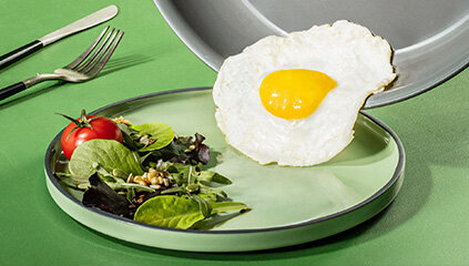 Na zielonym talerzu znajduje się sałatka z liści i pomidorków koktajlowych, obok której leży jajko sadzone. W tle widoczna jest patelnia, z której jajko zostało zsunięte.